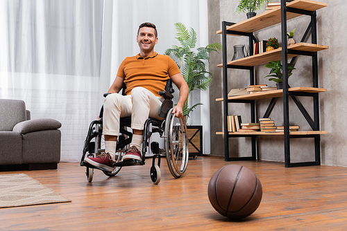 joyful man smiling at camera while sitting in wheelchair near basketball on floor