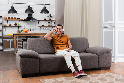 cheerful man listening music in headphones while sitting on couch in modern apartment