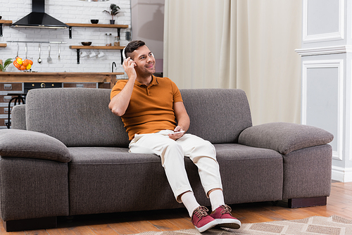 joyful man holding smartphone while listening music in headphones on sofa