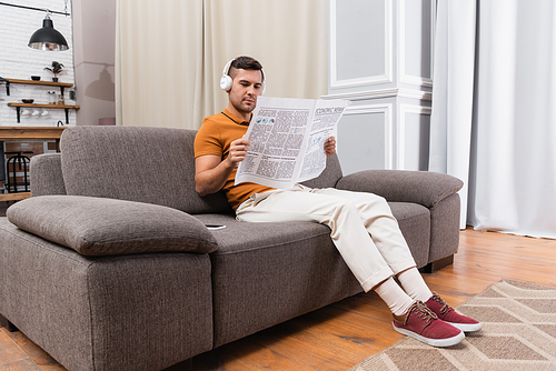 man in headphones listening music and reading newspaper on sofa at home