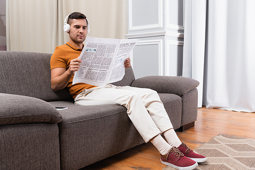 young man reading newspaper while listening music in headphones on couch at home