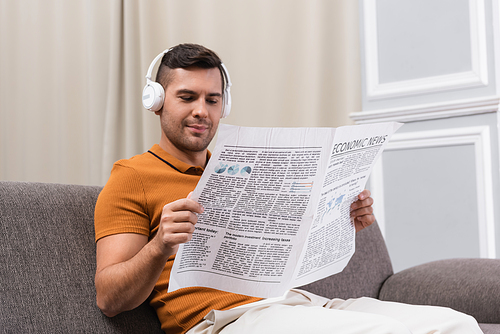 positive man in wireless headphones reading newspaper on sofa at home