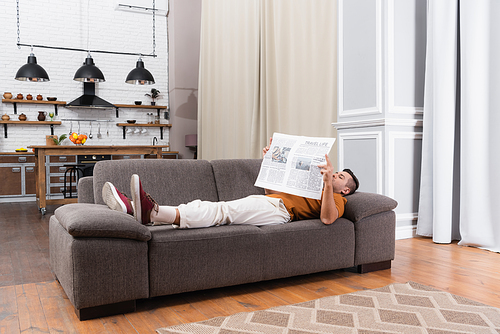 young man reading newspaper while lying on sofa in modern apartment