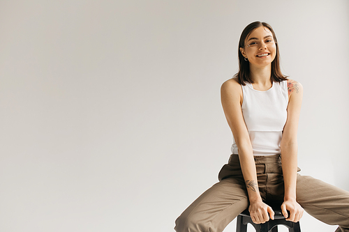young smiling woman in white top and beige trousers sitting on stool on grey background