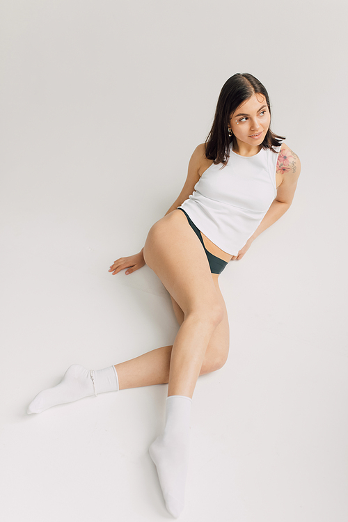 relaxed young woman in white top, socks and black panties lying on grey background