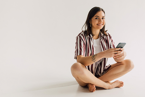 positive young woman in white top and striped shirt sitting with crossed legs and holding smartphone on grey background