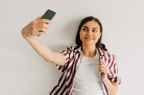 positive young woman in striped shirt taking selfie with smartphone on grey background