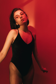 sensual young woman with bob haircut in black bodysuit and sunglasses on red background with shadows