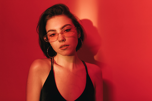 sensual woman with bob haircut in black bodysuit and sunglasses with half-opened mouth on red background with shadows