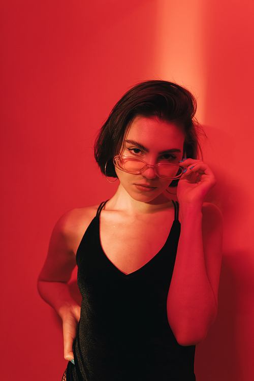 serious young woman with bob haircut in black bodysuit adjusting sunglasses and  on red background with shadows
