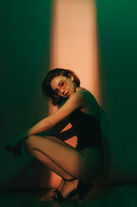 young woman sitting in black bodysuit and fishnet tights in squat pose on green and beige background