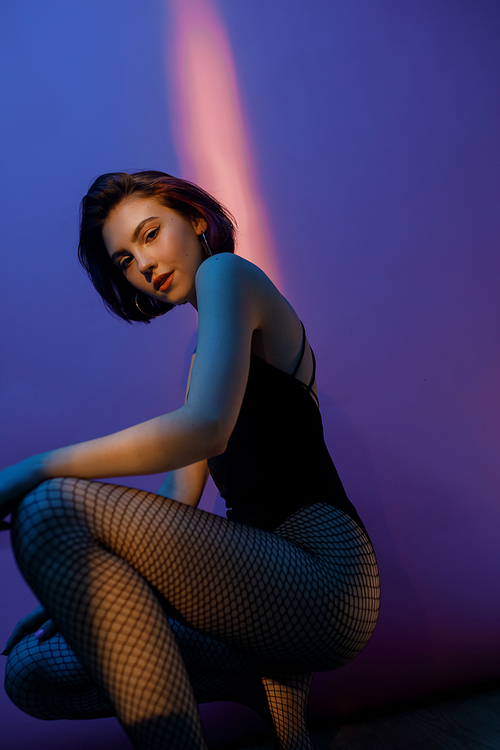 young woman sitting in black bodysuit and fishnet tights in pose on violet and pink background