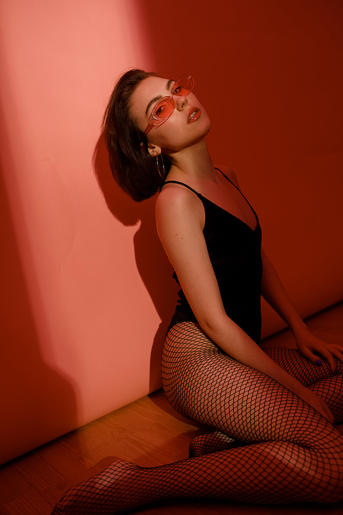 young woman sitting on floor in black bodysuit and fishnet tights on red background