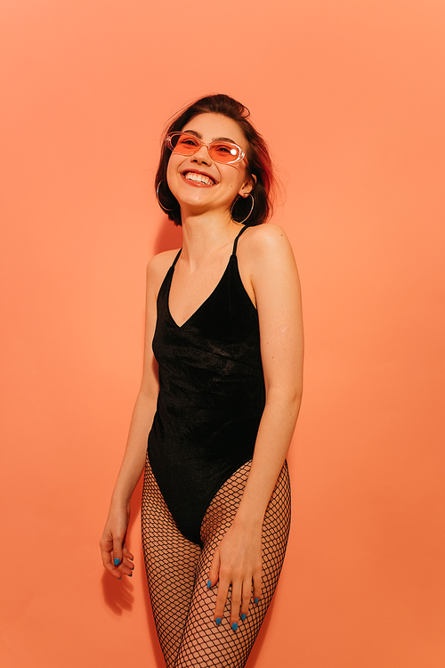 happy young woman in black bodysuit and sunglasses posing on orange background