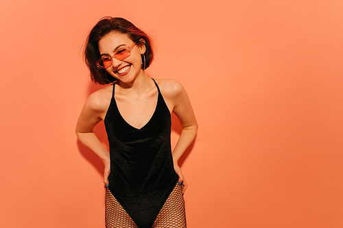 smiling young woman in sunglasses, black bodysuit and fishnet tights posing on orange background