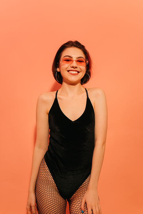 smiling young woman in black bodysuit and sunglasses posing on orange background