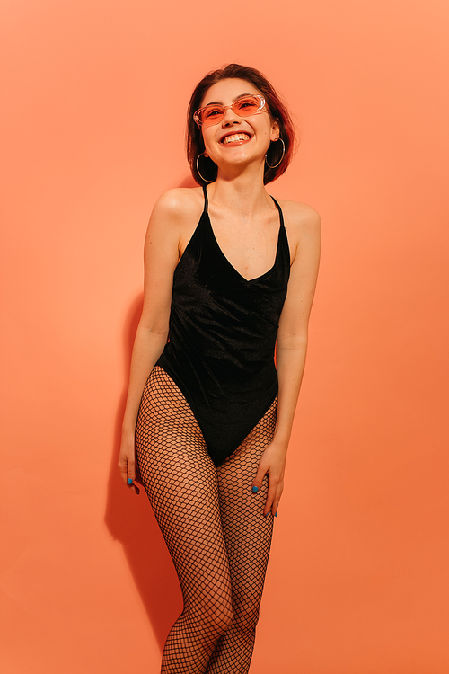 smiling young woman posing in black bodysuit and fishnet tights on orange background