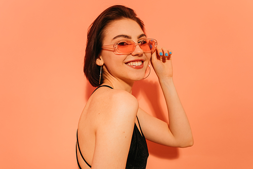 smiling young woman posing,  and adjusting sunglasses on orange background