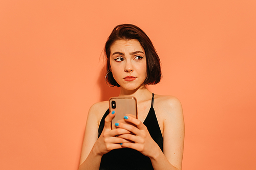 tense young woman with cellphone in hands in black tank top on orange background