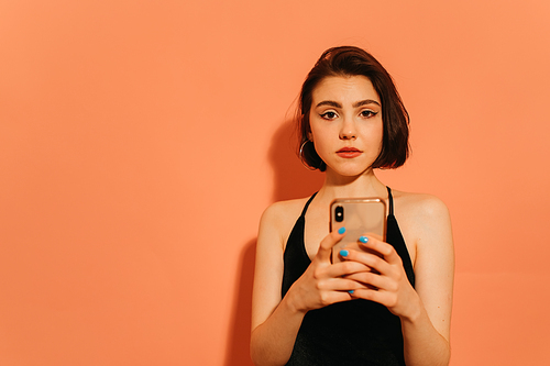 young adult woman holding smartphone in hands and tensely  on orange background