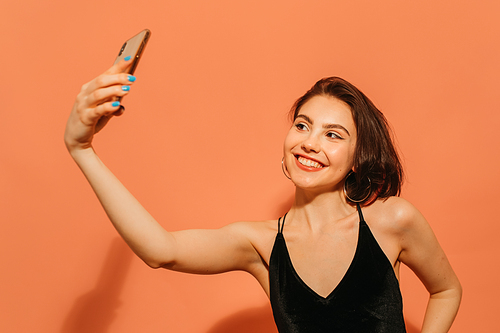 positive young woman taking selfie and smiling on orange background