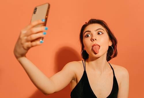 young woman taking selfie and grimacing with sticking out tongue on orange background
