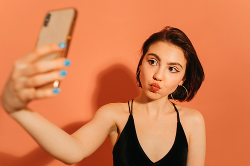 young woman taking selfie and grimacing with pouting lips on orange background
