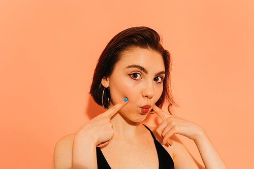 young woman  and grimacing with pouting lips and hands near face on orange background