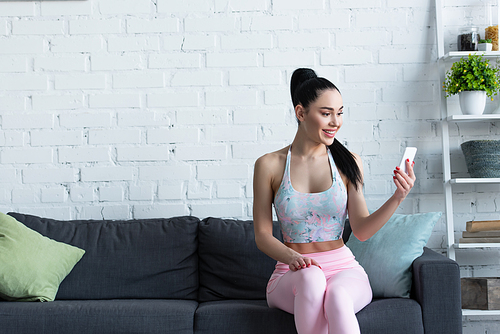 sportive woman smiling while sitting on couch and using smartphone