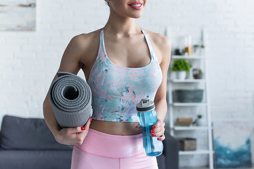 partial view of smiling woman holding fitness mat and sports bottle at home
