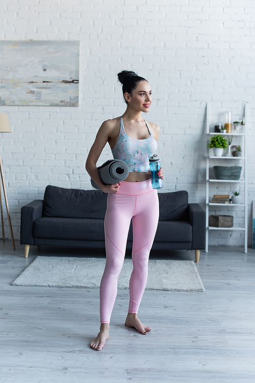 sportive barefoot woman standing with yoga mat and sports bottle at home