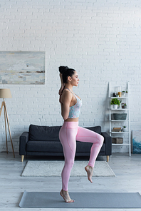 side view of young woman standing on one leg with hands behind back while practicing yoga at home