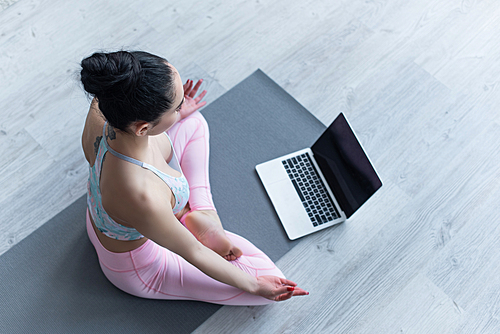 overhead view of woman meditating in lotus pose on yoga mat near laptop