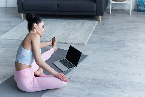 young woman looking at laptop while stretching on yoga mat at home