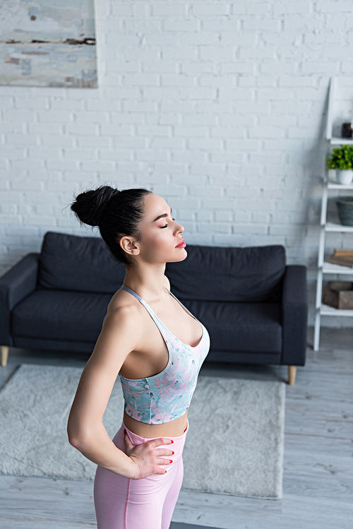 young woman in sports bra standing with hands on hips and closed eyes while training at home