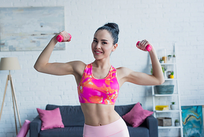 sportive woman smiling at camera while working out with dumbbells at home