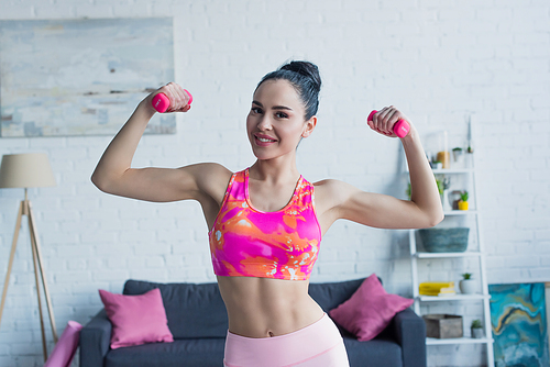sportive woman smiling at camera while working out with dumbbells at home