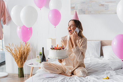 Excited woman talking on cellphone and holding birthday cake with candles on bed