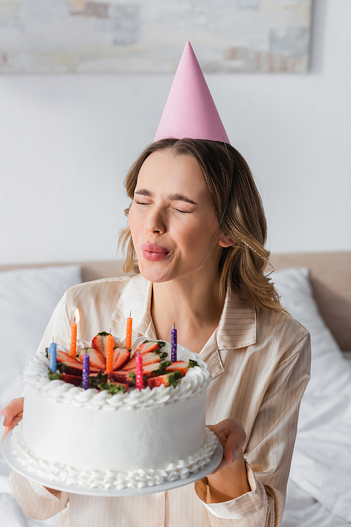 Woman in pajama and party cap blowing out candles on birthday cake