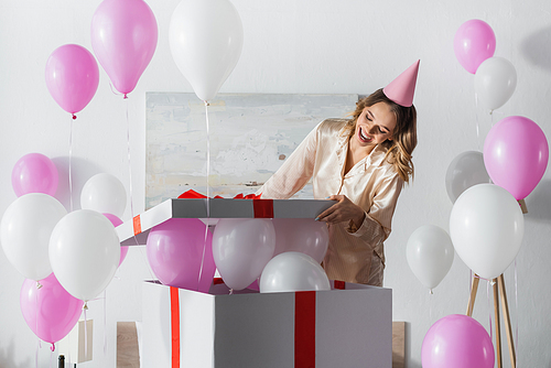 Woman in party cap and pajama opening gift box with festive balloons at home
