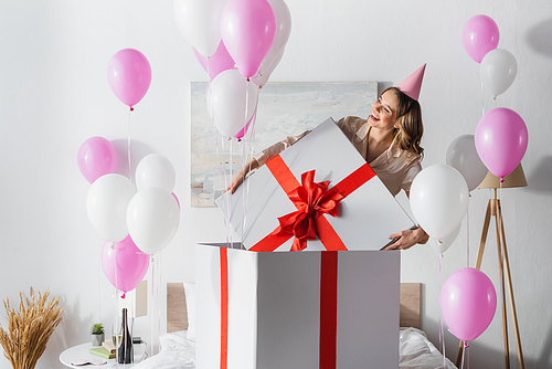 Smiling woman holding cap of huge present near festive balloons in bedroom
