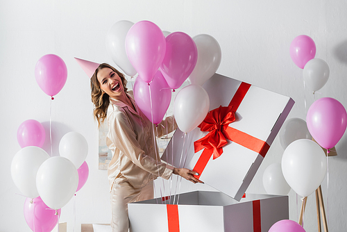 Positive woman standing near big gift box with balloons during birthday celebration at home