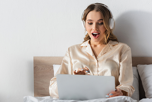 Excited woman in headphones looking at blurred laptop on bed