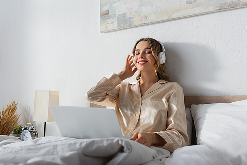 Cheerful woman listening music in headphones near blurred laptop on bed