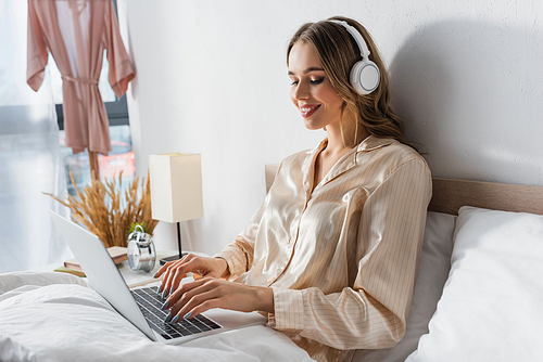 Smiling freelancer in headphones using laptop in bedroom