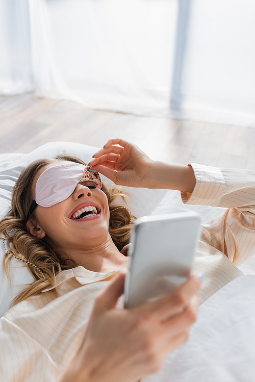 Cheerful woman in sleeping mask using smartphone on bed