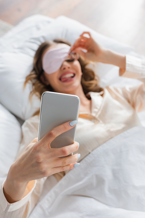 Smartphone in hand of blurred woman in sleeping mask on bed