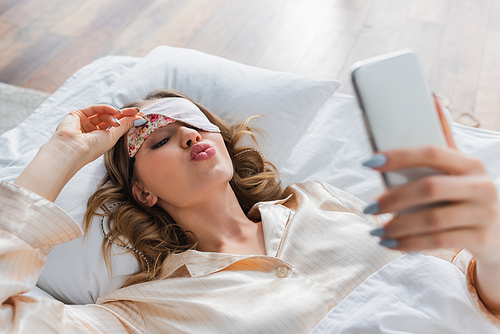 Woman in sleeping mask pouting lips and using smartphone on bed