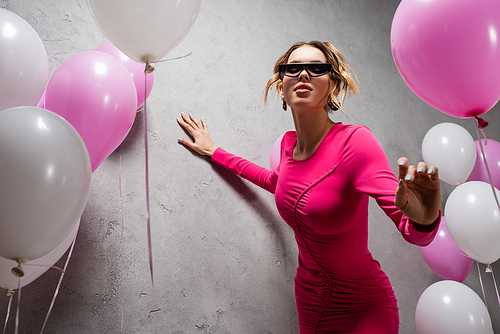 Low angle view of stylish woman in sunglasses and dress near festive balloons and concrete wall