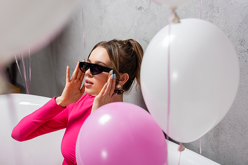 Young woman in sunglasses sitting in bathtub near blurred balloons on foreground